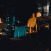 A moody, shadow-filled cafe scene with colorful chairs and tables in dramatic lighting.