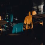 A moody, shadow-filled cafe scene with colorful chairs and tables in dramatic lighting.