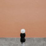 A minimalistic photo of a person in a hat against a large orange wall, perfect for design needs.