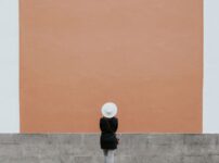 A minimalistic photo of a person in a hat against a large orange wall, perfect for design needs.