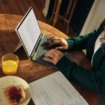Woman typing on laptop at wooden table with breakfast.